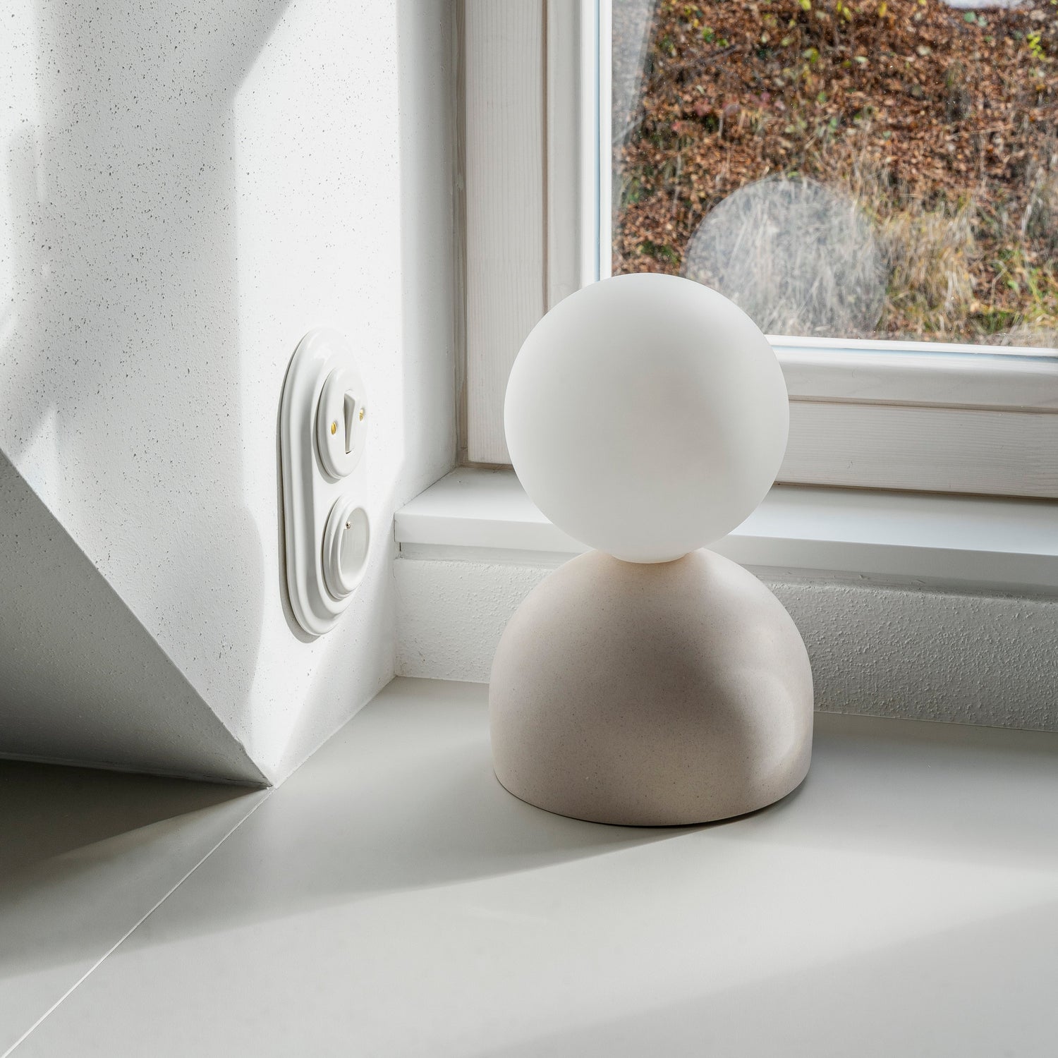 NUUK table lamp with a matte concrete base and frosted glass globe shade, placed on a white windowsill with natural light casting soft shadows on the wall behind it.